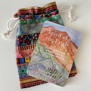 May include: A card with a watercolor painting of a mountain range with green foliage and a blue sky. The card has the text 'Spirit of the Earth' in white lettering. The text below the image reads 'Empowering Affirmations Inspired by Nature for Self-Reflection and Growth' and 'Designed & Illustrated by Michelle Pugh'. The card deck is laid on top of a colorful drawstring bag that is used to hold the deck when not in use.