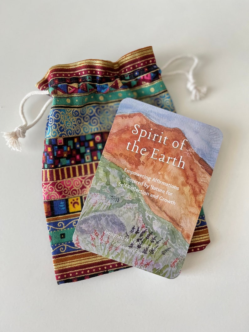 May include: A card with a watercolor painting of a mountain range with green foliage and a blue sky. The card has the text 'Spirit of the Earth' in white lettering. The text below the image reads 'Empowering Affirmations Inspired by Nature for Self-Reflection and Growth' and 'Designed & Illustrated by Michelle Pugh'. The card deck is laid on top of a colorful drawstring bag that is used to hold the deck when not in use.