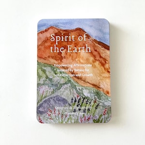 May include: A card with a watercolor illustration of a mountain range with a blue sky. The text "Spirit of the Earth" is written in white on the card. The text "Empowering Affirmations Inspired by Nature for Self-Reflection and Growth" is written in black below the title. The text "Designed & Illustrated by Michelle Pugh" is written in black at the bottom of the card.