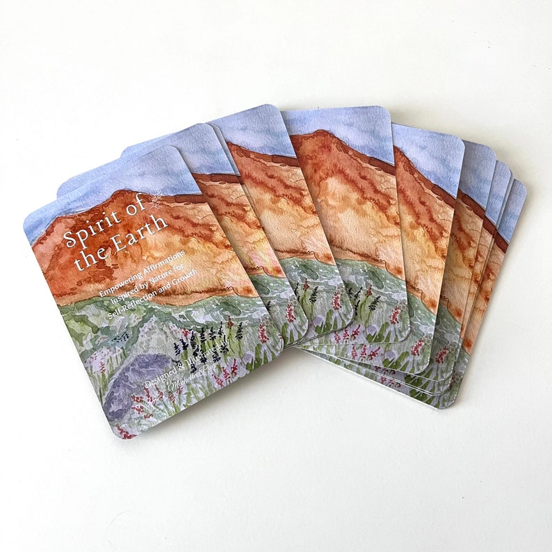May include: A set of affirmation cards with a watercolor illustration of a landscape with a mountain range, green grass, and flowers. The cards are titled "Spirit of the Earth" and feature the text "Empowering Affirmations Inspired by Nature for Self-Reflection and Growth".