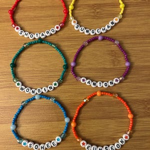May include: Six beaded bracelets in various colors: red, yellow, green, purple, blue, and orange. Each bracelet features white letter beads spelling out names: SHOTARO, SUNGCHAN, WONBIN, ANTON, SOHEE, and EUNSEOK. The bracelets are on a wooden surface.