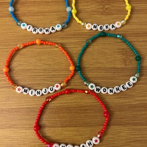 May include: Five beaded bracelets in various colors: blue, yellow, orange, green, and red. Each bracelet features a name spelled out with white letter beads. The bracelets are on a wooden surface.