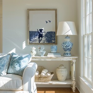 Pode incluir: Cena de sala de estar com sof&aacute; branco e almofadas florais azuis. Uma consola branca desgastada exibe uma obra de arte emoldurada de um c&atilde;o, uma l&acirc;mpada azul e branca e objetos decorativos. Tema costeiro.