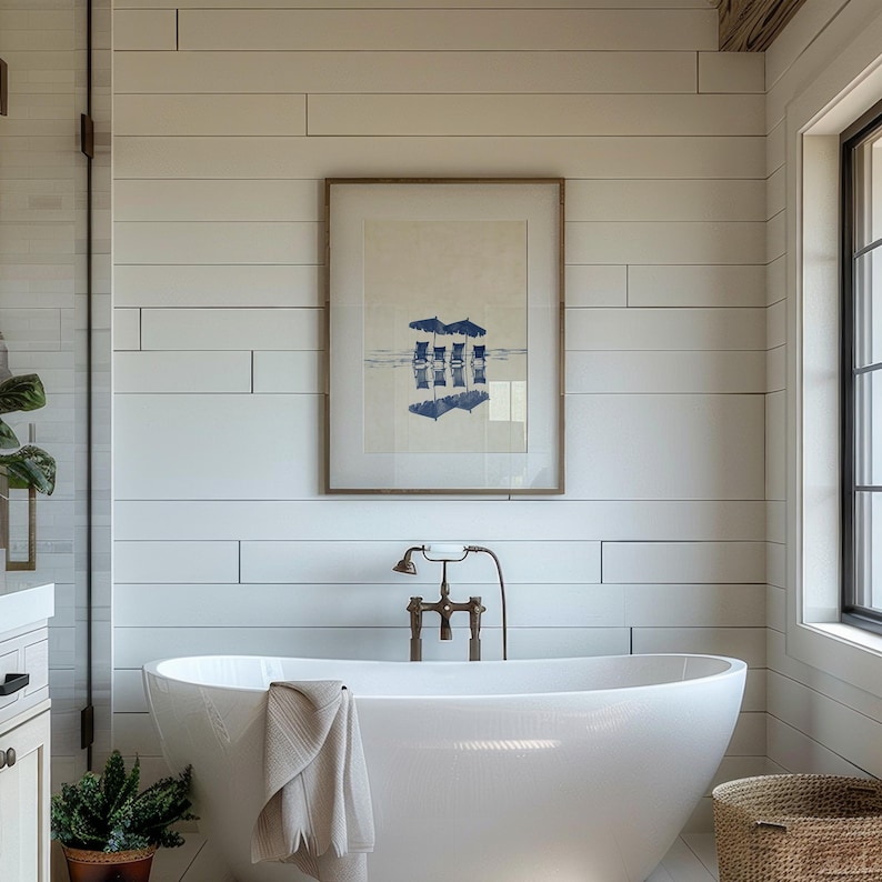 May include: A white freestanding bathtub with a white towel draped over the edge. The bathtub is in a bathroom with white shiplap walls, a framed print of beach chairs under umbrellas, and a brass faucet.