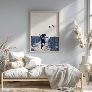 Pode incluir: Impress&atilde;o emoldurada com um c&atilde;o preto correndo por um campo, com dois p&aacute;ssaros no c&eacute;u. A obra de arte &eacute; em tons de azul e branco, com uma moldura de madeira natural. A impress&atilde;o est&aacute; acima de uma cama.