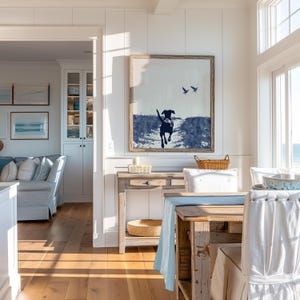 Pode incluir: Uma obra de arte emoldurada retrata um c&atilde;o correndo em tons de azul e branco, com dois p&aacute;ssaros no c&eacute;u. A obra est&aacute; pendurada acima de uma mesa de console de madeira com uma cesta de vime e uma vela. A sala de jantar inclui uma mesa com um corredor azul e cadeiras brancas.