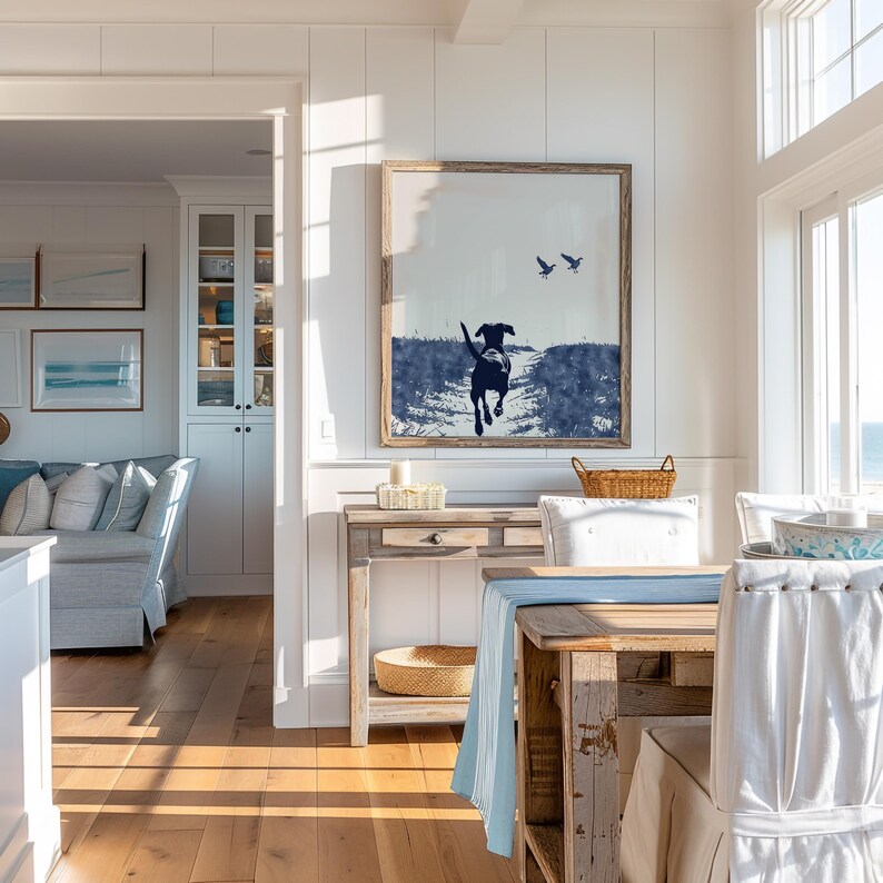 Pode incluir: Uma obra de arte emoldurada retrata um c&atilde;o correndo em tons de azul e branco, com dois p&aacute;ssaros no c&eacute;u. A obra est&aacute; pendurada acima de uma mesa de console de madeira com uma cesta de vime e uma vela. A sala de jantar inclui uma mesa com um corredor azul e cadeiras brancas.