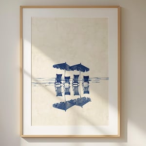 May include: A framed print of a beach scene with four blue chairs and umbrellas reflected in the water. The print is on a beige background.