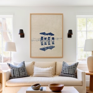 May include: A framed print of a beach scene with four blue lounge chairs under three blue umbrellas reflected in the water. The print is hanging above a beige sofa with two patterned pillows and a white throw pillow.