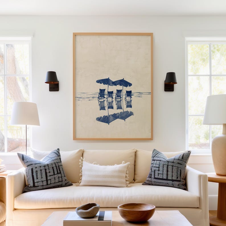 May include: A framed print of a beach scene with four blue lounge chairs under three blue umbrellas reflected in the water. The print is hanging above a beige sofa with two patterned pillows and a white throw pillow.