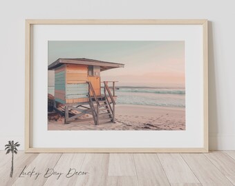 Könnte beinhalten: Gerahmter Druck mit einer Strandwache an einem Sandstrand. Die Station ist in Pastelltönen gehalten, mit einer Holztreppe zum Eingang. Im Hintergrund sind das Meer und der Himmel zu sehen, was eine ruhige Küstenlandschaft schafft.