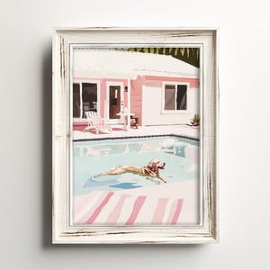 Può includere: Stampa artistica incorniciata raffigurante un cane che nuota in una piscina. L'opera presenta una casa rosa, una piscina con acqua azzurra e un cane con pelo marrone. La cornice è bianca con finitura anticata. L'opera è un'illustrazione colorata.