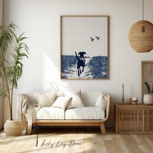 Pode incluir: Uma impress&atilde;o emoldurada com uma ilustra&ccedil;&atilde;o em estilo aquarela de um c&atilde;o correndo em um caminho, com dois p&aacute;ssaros no c&eacute;u. A obra de arte &eacute; principalmente em tons de azul e branco, com uma moldura de madeira. A impress&atilde;o &eacute; exibida acima de um sof&aacute; branco.