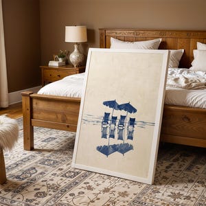 May include: A white framed print of a blue and white watercolor painting of four chairs with umbrellas on a beach. The print is leaning against a wooden bed frame in a bedroom.