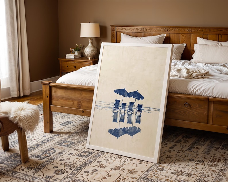 May include: A white framed print of a blue and white watercolor painting of four chairs with umbrellas on a beach. The print is leaning against a wooden bed frame in a bedroom.
