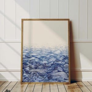 Può includere: Una stampa incorniciata di un design astratto di onde oceaniche blu e bianche. Le onde sono composte da linee sottili che creano un effetto testurizzato. La stampa è in una cornice di legno chiaro ed è appesa a una parete bianca.
