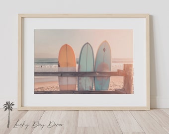 May include: Three surfboards leaning against a wooden railing on a beach. The surfboards are orange, blue, and green. The beach is sandy and the sky is a soft pink and orange.