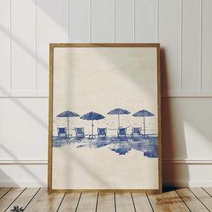 May include: A watercolor painting of a beach scene with blue umbrellas and chairs. The chairs are facing the ocean and the umbrellas are open. The painting is in a light brown frame.