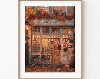 May include: A framed print of a cozy bookstore facade. The shop, with the text "the book shop" above the windows, is framed by autumn foliage and a vintage bicycle. A warm glow emanates from the interior, suggesting a welcoming atmosphere.