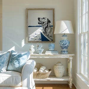 May include: A white, distressed, wooden console table with two shelves. The top shelf has a white lamp with a white shade, a blue and white porcelain vase, and a book. The bottom shelf has a white wicker basket with white balls, and a white ceramic jar with a decorative pattern. A framed black and white print of a dog in a boat hangs on the wall above the table.