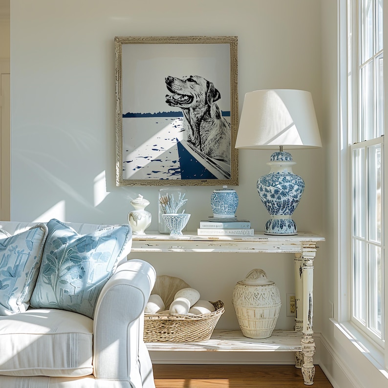 May include: A white, distressed, wooden console table with two shelves. The top shelf has a white lamp with a white shade, a blue and white porcelain vase, and a book. The bottom shelf has a white wicker basket with white balls, and a white ceramic jar with a decorative pattern. A framed black and white print of a dog in a boat hangs on the wall above the table.