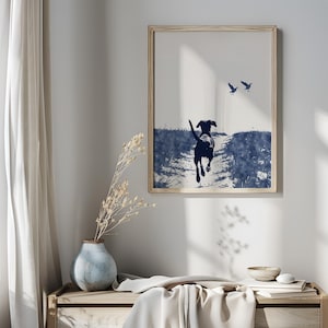 Pode incluir: Impress&atilde;o emoldurada com uma silhueta azul marinho de um c&atilde;o correndo em um caminho, com dois p&aacute;ssaros voando acima. A obra de arte tem um efeito aquarela, em um fundo claro. A moldura &eacute; de cor de madeira clara.