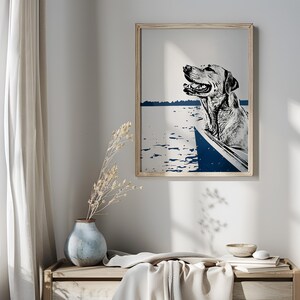 May include: A black and white illustration of a dog in a boat on a blue and white lake. The dog is looking to the right and has its mouth open. The illustration is framed in a light wood frame.