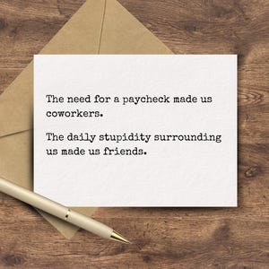 May include: A white rectangular greeting card with black typewriter-style text that reads: The need for a paycheck made us coworkers. The daily stupidity surrounding us made us friends. The card is placed next to a tan envelope and a gold pen.