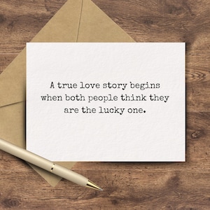 May include: A white rectangular greeting card featuring the black printed text: A true love story begins when both people think they are the lucky one. The card is positioned over a brown paper envelope with a gold-toned ballpoint pen nearby.