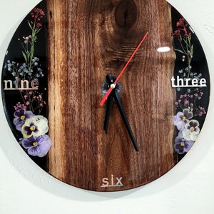 May include: A round wooden clock with a black background and floral accents. The clock face has Roman numerals and the words "twelve", "three", "nine", and "six" printed on it. The clock hands are black and red.