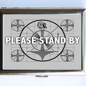 May include: A silver metal cigarette case with a black and white image of a vintage television test pattern. The text "PLEASE STAND BY" is printed in black letters on the case.