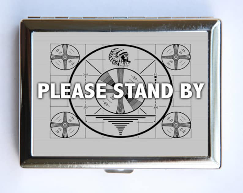 Puede incluir: Una cajetilla de cigarrillos de metal plateado con una imagen en blanco y negro de un patr&oacute;n de prueba de televisi&oacute;n vintage. El texto "PLEASE STAND BY" est&aacute; impreso en letras negras en la cajetilla.