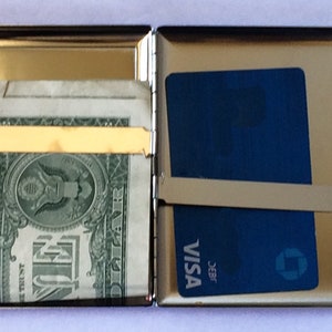 May include: A silver metal cigarette case with a gold trim. The case is open and has a dollar bill and a credit card inside. The credit card is a Visa card with the text "J.E.B.R. VISA" printed on it.