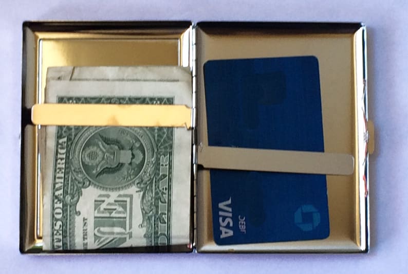May include: A silver metal cigarette case with a gold trim. The case is open and has a dollar bill and a credit card inside. The credit card is a Visa card with the text "J.E.B.R. VISA" printed on it.