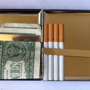 May include: A silver cigarette case with a gold-colored metal band and a hinged lid. The case is open and contains a pack of cigarettes and a few dollar bills.