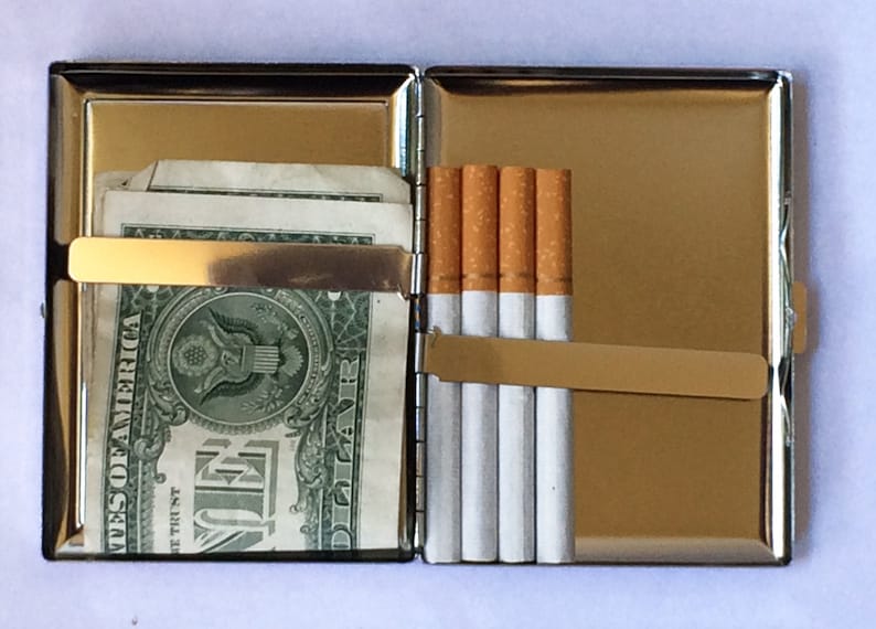 May include: A silver cigarette case with a gold-colored metal band and a hinged lid. The case is open and contains a pack of cigarettes and a few dollar bills.