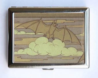 May include: A silver metal compact with a graphic of a brown bat flying over a green and brown cloud. The bat has a long snout and large ears.
