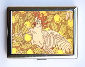 May include: A vintage-style compact with a colorful illustration of a white bird with a red and orange crest, surrounded by yellow lemons and green leaves on a brown background.