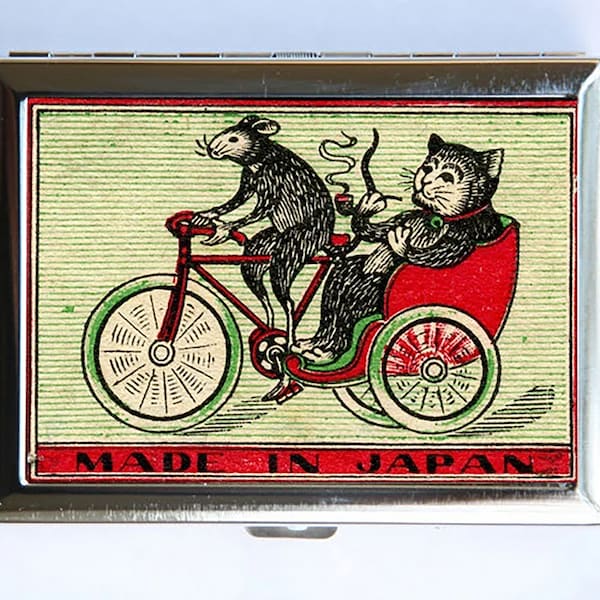 May include: A vintage metal matchbox featuring a black cat and a mouse riding a bicycle. The cat is in a red carriage with a green frame. The image has a green and white striped background with the text "MADE IN JAPAN".