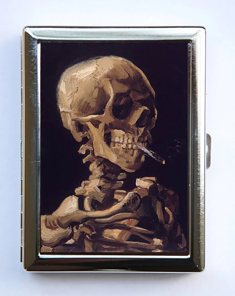 May include: A metal cigarette case with a printed image of a skull smoking a cigarette. The skull is painted in a realistic style with a dark background.