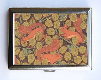 May include: A silver metal compact with a colorful design featuring three red squirrels in a leafy pattern. The squirrels are surrounded by green and brown leaves.