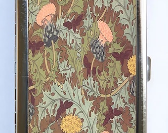 May include: A silver cigarette case with a floral design featuring pink, yellow, and brown flowers on a green background.