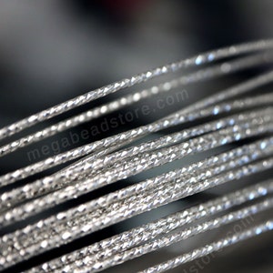 May include: Close-up of silver-colored, braided wire with a diamond-shaped pattern. The wire is coiled and has a shiny, metallic finish.