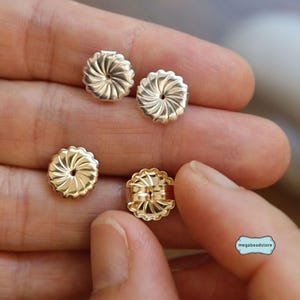 May include: Four gold-toned earring backs. Two are flat and two are hinged. The backs have a swirl design.