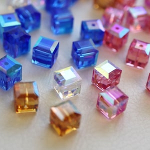 May include: A collection of small, faceted cube-shaped beads in various translucent colors including blue, pink, and amber. Each bead features a central hole and a reflective, iridescent finish that catches the light, creating a sparkling effect.