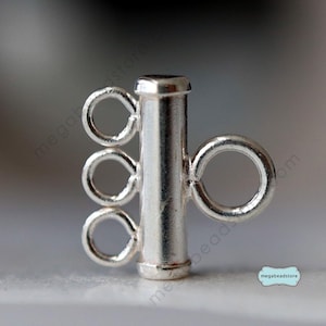 Puede incluir: Cierre de metal plateado con tres bucles para sujetar múltiples hebras de cuentas o cadenas. El cierre tiene una forma cilíndrica y un acabado liso.