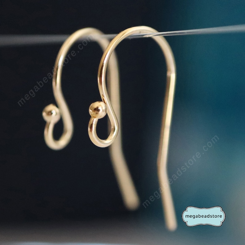 May include: Close-up of two gold-colored earring hooks. Each hook has a curved design with a small ball at the bottom and a loop. The hooks are suspended against a dark blue background. The text "megabeadstore.com" is visible.