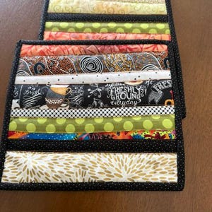 May include: Two handmade fabric coasters with a patchwork design. Each coaster features horizontal strips of various patterned fabrics, including floral, polka dots, and coffee-themed prints. The edges are finished with a black dotted border.