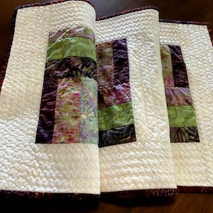 May include: A white quilted table runner with a burgundy border and a patchwork design of green, purple, and brown fabric squares.