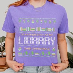May include: A purple t-shirt with a Christmas-themed design featuring a green and white Christmas tree, a green and white ornament, and the text "I'd rather stay at my library this Christmas".
